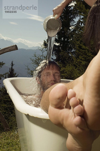 Der junge Mann  der in der Badewanne lag  schüttet das Wasser auf den Mann  und zwar auf den Mann.