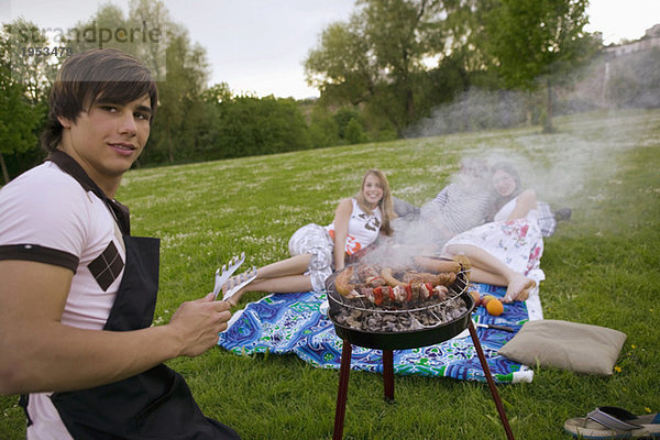 Junger Mann beim Grillen junger Frauen im Hintergrund