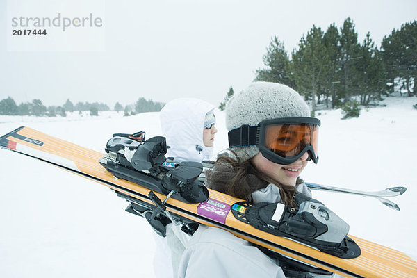 Zwei junge Skifahrer  die zusammen laufen und Skier auf den Schultern tragen.