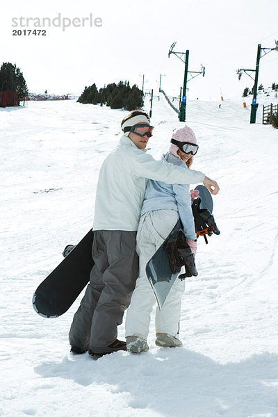 Zwei junge Snowboarder gehen zusammen  schauen über die Schultern in die Kamera.