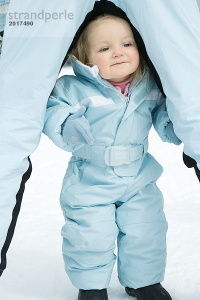 Kleinkind zwischen Geschwisterbeinen stehend  im Skianzug gekleidet  lächelnd