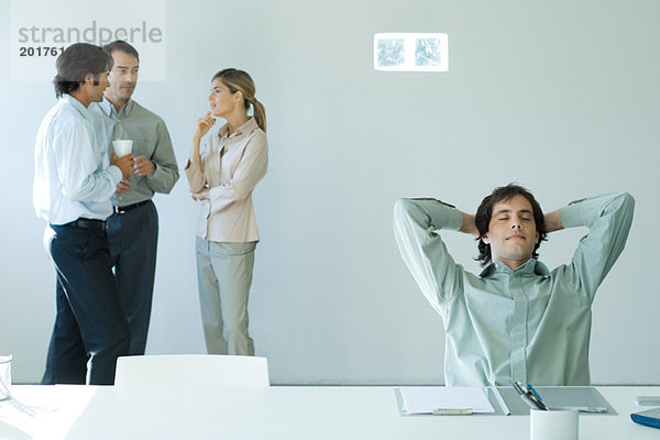Geschäftsmann am Schreibtisch mit geschlossenen Händen hinter Kopf und Augen  Kollegen im Hintergrund