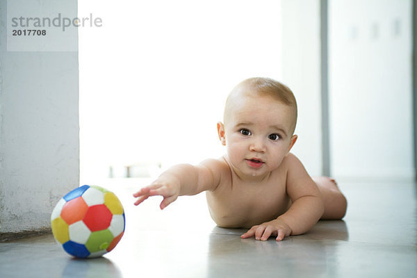 Baby liegt auf dem Boden  greift nach dem Ball und schaut in die Kamera.