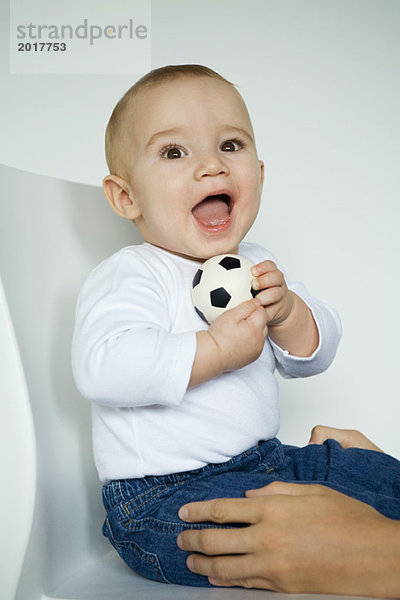 Baby sitzt im Stuhl und hält den Ball  die Hände der Mutter halten die Beine des Babys.