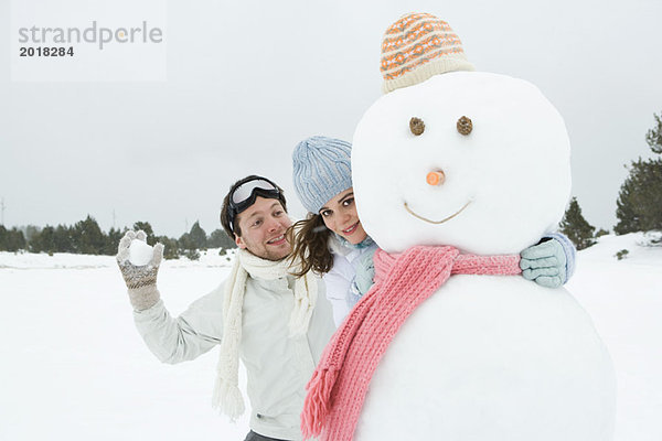 Weibchen umarmt Schneemann und lächelt in die Kamera  Freund steht dahinter  hält Schneeball in der Hand