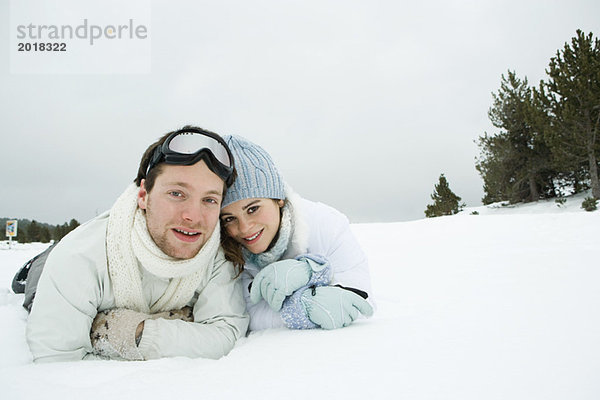 Junges Paar im Schnee nebeneinander liegend  lächelnd vor der Kamera  Porträt