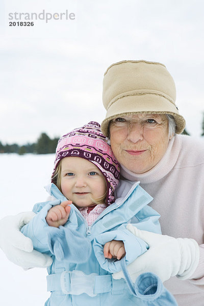 Seniorin umarmt Kleinkind Mädchen  beide lächelnd  in Winterkleidung gekleidet  Portrait