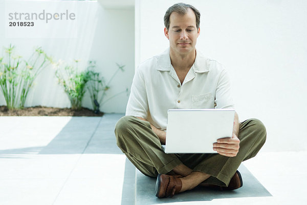 Mann am Boden sitzend mit Laptop  lächelnd  volle Länge