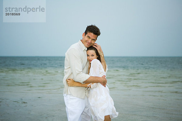 Mann und junge Begleiterin am Strand  umarmend  in die Kamera schauend