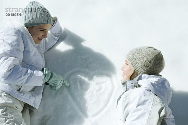 Teenagermädchen auf Schnee liegend  lachend  Herz mit Initialen auf Schneeoberfläche gezeichnet