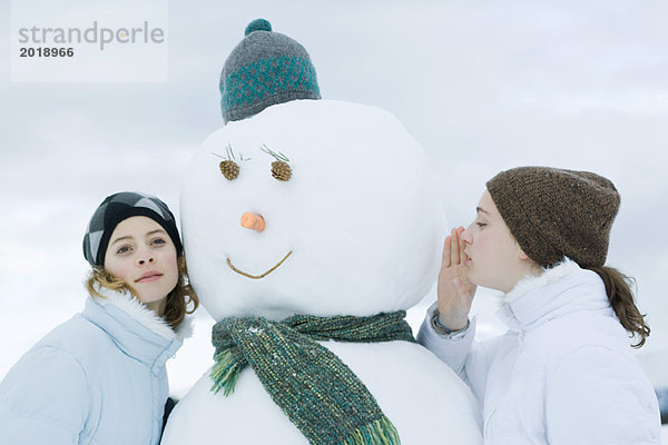 Junge Mädchen auf beiden Seiten des Schneemanns  eine flüstert  die andere hört zu.