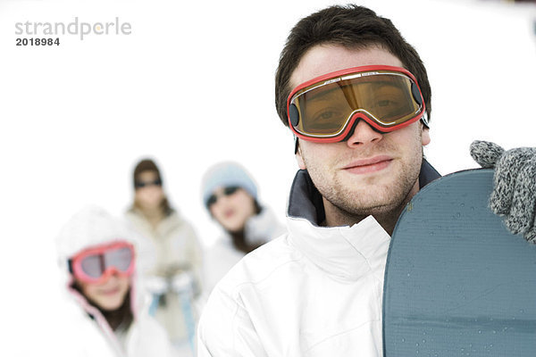 Junger Mann stehend mit Snowboard  Freunde im Hintergrund  Portrait