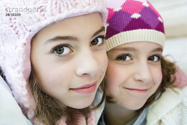 Zwei junge Schwestern lächeln vor der Kamera  beide mit Strickmützen  Wange an Wange  Portrait