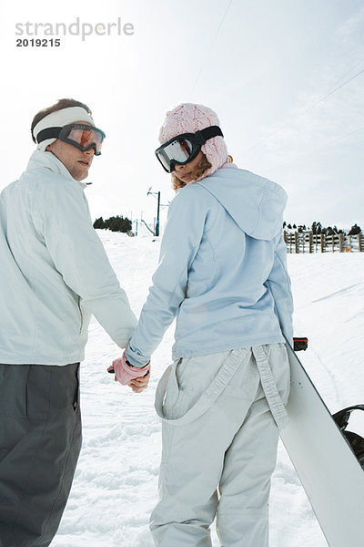 Zwei junge Freunde gehen  Händchen halten  über die Schultern schauen  einer trägt Snowboard  Rückansicht