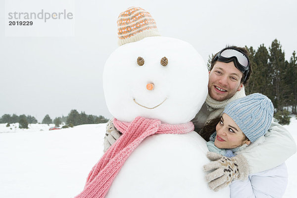 Zwei junge Freunde stehen mit Schneemann  beide lächelnd  einer schaut in die Kamera.