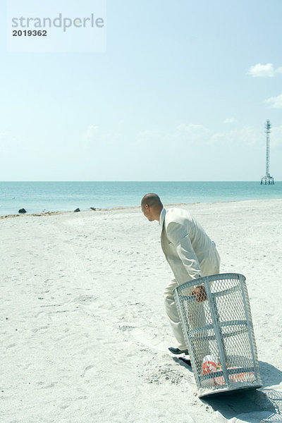 Am Strand  Mann im Anzug zieht Mülltonne zum Meer  Seitenansicht  volle Länge