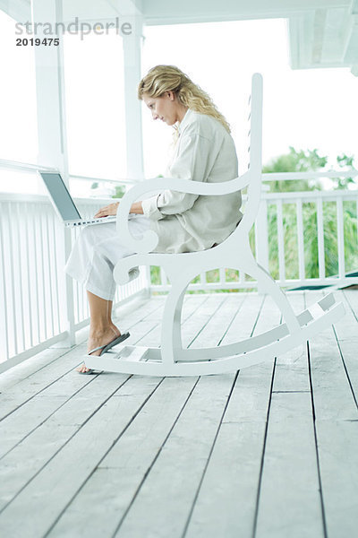 Frau im Schaukelstuhl auf der Veranda sitzend  mit Laptop  volle Länge