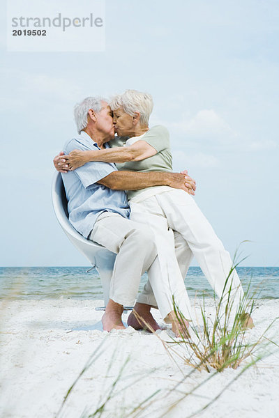Seniorenpaar sitzt zusammen im Stuhl am Strand  umarmt und küsst sich  Frau sitzt auf dem Schoß des Mannes  volle Länge