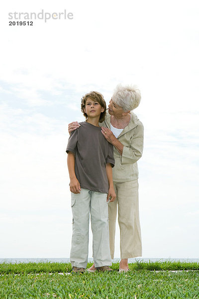 Seniorin und Enkelin auf Rasen stehend  Frau im Gespräch mit dem Jungen über die Schulter  volle Länge