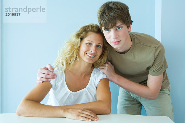 Jugendlicher Junge neben Mutter am Tisch sitzend  Wange an Wange  Kamera lächelnd  Porträt