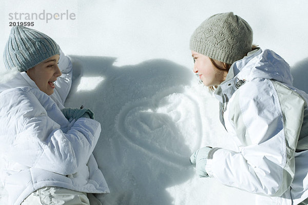 Zwei Teenagermädchen auf Schnee liegend  auf das Herz schauend  mit Initialen im Schnee gezeichnet