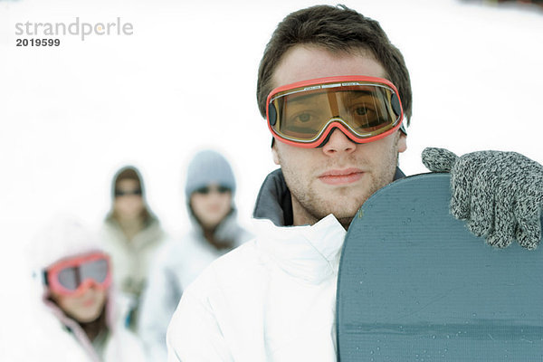 Junger Snowboarder mit Blick auf die Kamera  Freunde im Hintergrund  Porträt