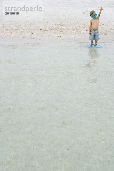 Kleiner Junge im flachen Wasser stehend  mit Schnorchelmaske und Flossen  ein Arm hochgezogen  volle Länge  Vorderansicht