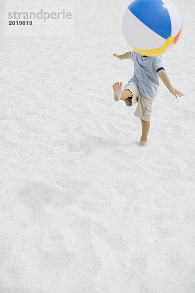 Junge tritt Strandball am Strand  Kopf durch Ball versteckt  hohe Winkelansicht  volle Länge