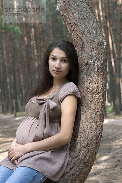 Eine schwangere Frau  die sich im Wald an einen Baum lehnt.