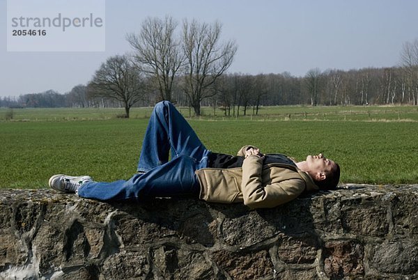 Ein Mann  der auf einer Steinmauer bei einem Feld liegt.