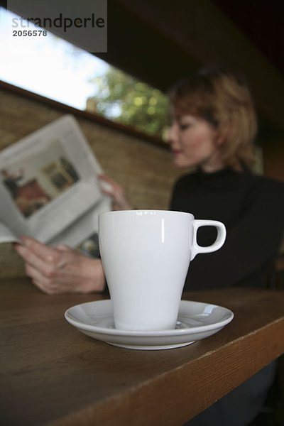 Eine Kaffeetasse auf einem Tisch im Café mit einer Zeitung lesenden Frau im Hintergrund