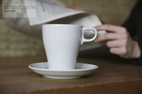 Eine Kaffeetasse auf einem Tisch im Café mit einer Zeitung lesenden Frau im Hintergrund