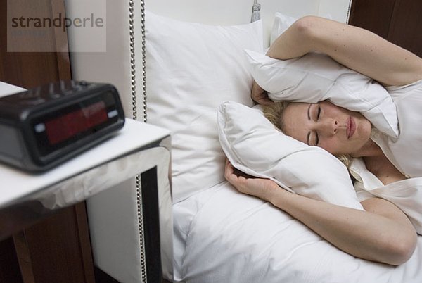 Eine Frau ignoriert im Bett liegend den Wecker  indem sie sich mit dem Kopfkissen die Ohren zuhält