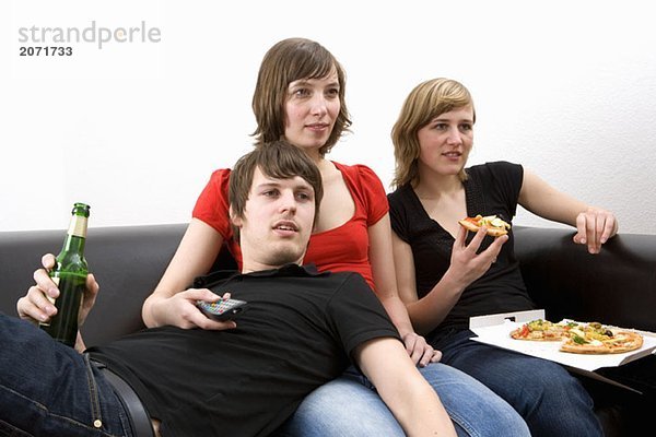 Drei junge Erwachsene sitzen gemeinsam auf dem Sofa  trinken Bier  essen Pizza und schauen fern.