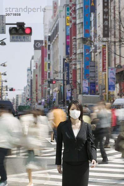 Eine Geschäftsfrau mit Mundschutz steht auf einem Zebrastreifen in der belebten Innenstadt  Tokio  Japan
