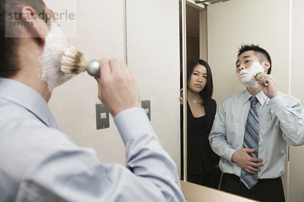 Ein Geschäftsmann steht mit Rasierschaum im Gesicht im Bad vor dem Spiegel  während er von einer hinter ihm stehenden Frau beobachtet wird
