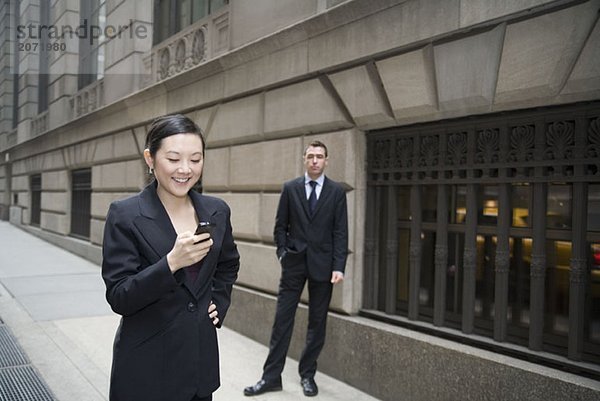 Eine Geschäftsfrau telefoniert mit einem Mobiltelefon während im Hintergrund ein Geschäftsmann wartet