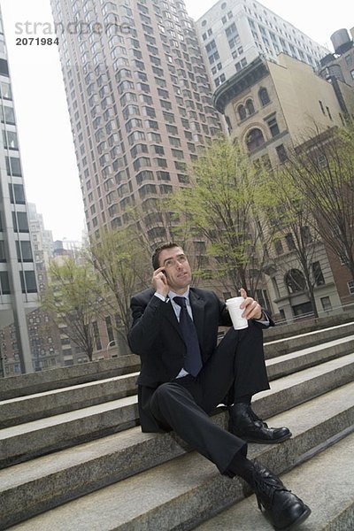 Ein Geschäftsmann sitzt auf einer Treppe und telefoniert einen Kaffeebecher haltend