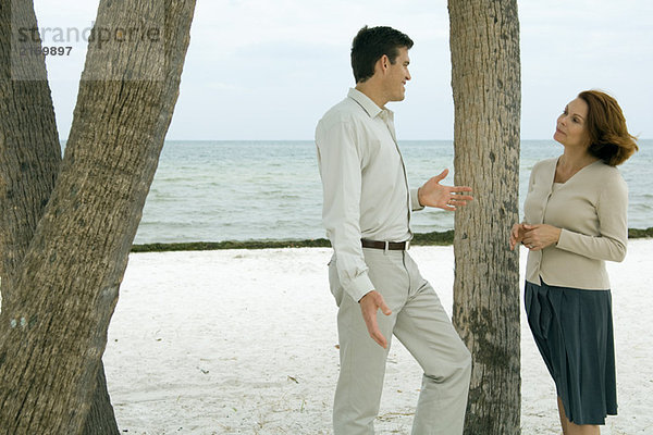 Mann und Frau stehen neben einem Baumstamm am Strand und lächeln sich an.