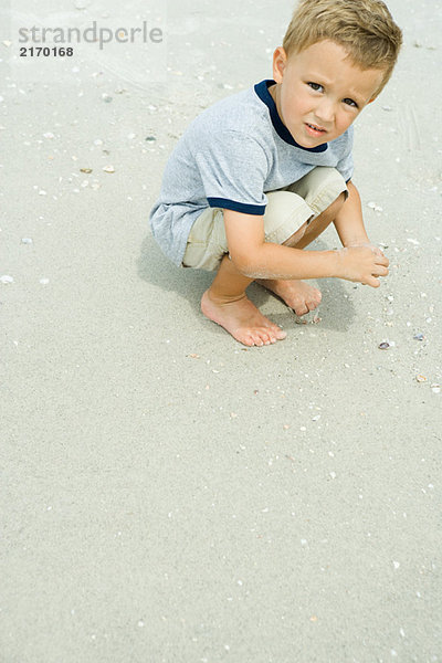 Junge hockt im Sand  schaut in die Kamera.