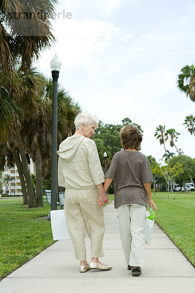 Junge und Großmutter gehen auf dem Bürgersteig  tragen Einkaufstaschen  reden  Rückansicht