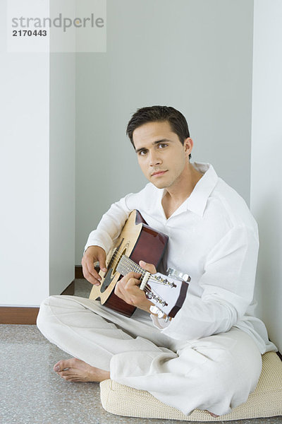 Junger Mann sitzt auf einem Kissen und spielt Akustikgitarre.