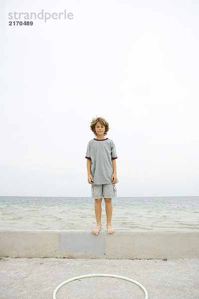 Junge steht auf einer niedrigen Wand am Strand und schaut in die Kamera.