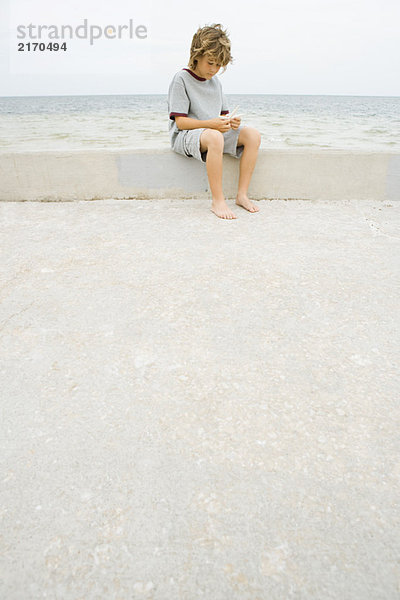 Junge sitzt auf einer niedrigen Wand am Strand und schaut auf den Seestern hinunter.