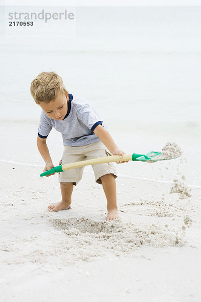 Kleiner Junge am Strand stehend  mit Schaufel im Sand grabend  nach unten schauend