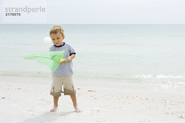 Kleiner Junge  der am Strand steht und versucht  den Ball mit dem Netz zu fangen.