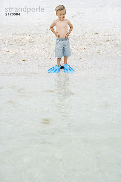 Kleiner Junge mit Flossen am Strand  am Wasser stehend  nach unten schauend