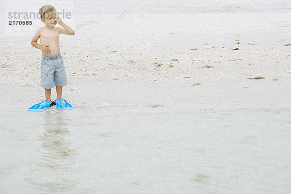 Kleiner Junge mit Flossen am Strand  steht am Wasserrand  reibt sich die Augen