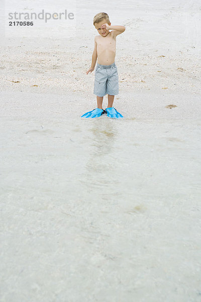Junge steht am Strand mit Flossen  reibt das Auge  schaut in die Kamera.