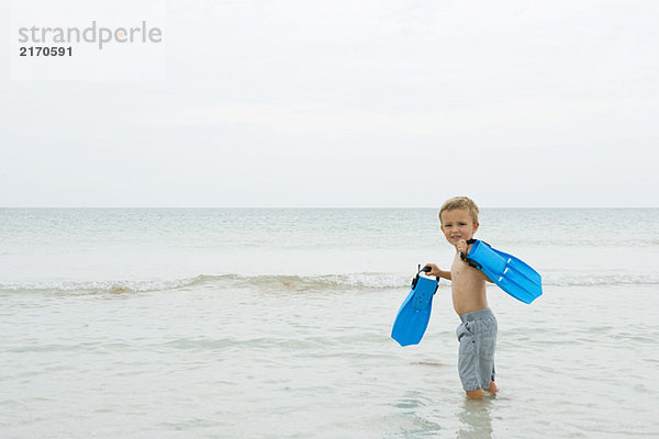 Junge steht knietief im Wasser  trägt Flossen und lächelt über die Schulter.